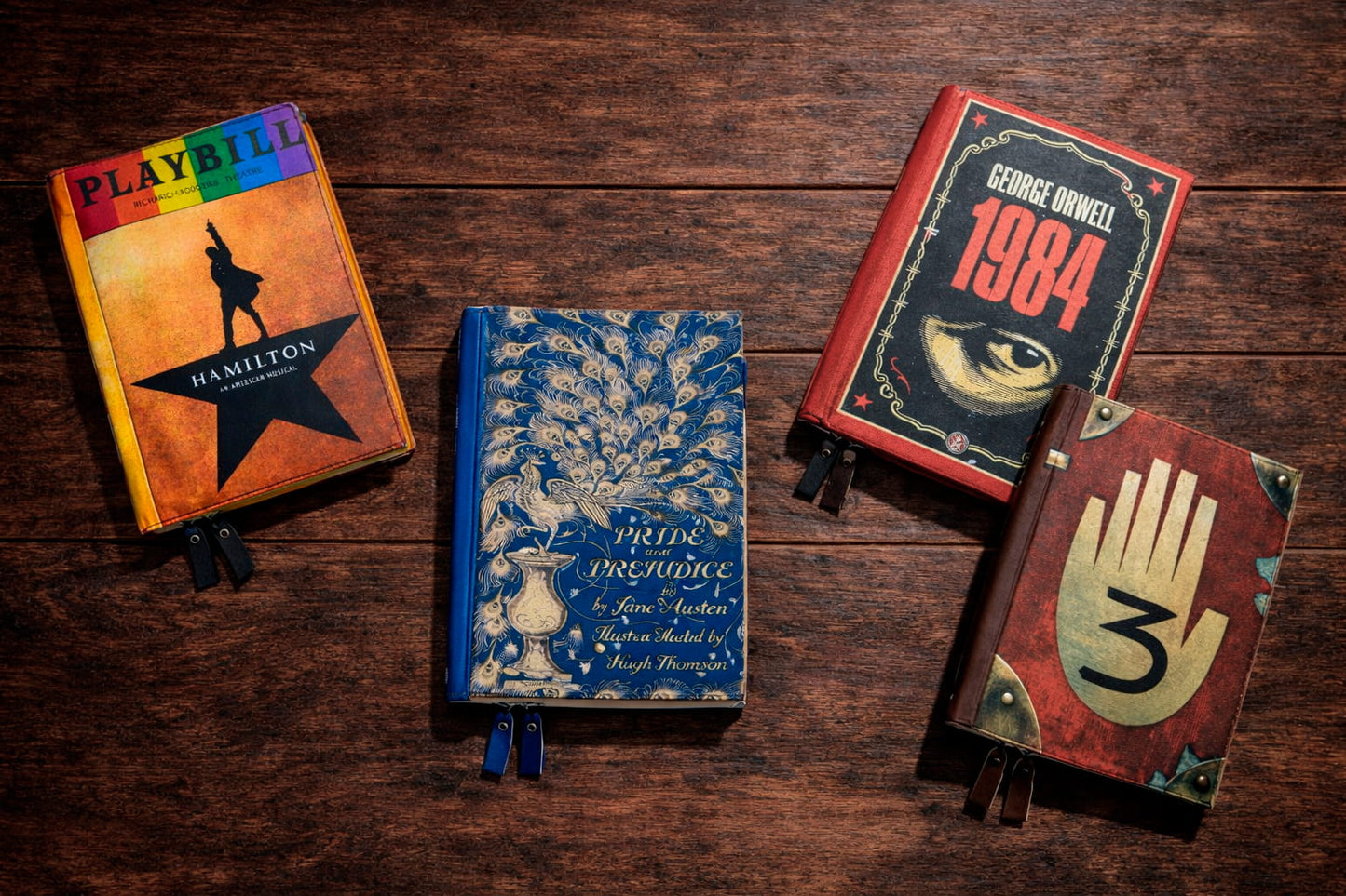 Four book-style cases for Kobo Libra Colour arranged in a scattered layout on a dark wooden table. The designs include a Hamilton Playbill cover, a blue Pride and Prejudice case with a peacock illustration, a 1984 by George Orwell cover